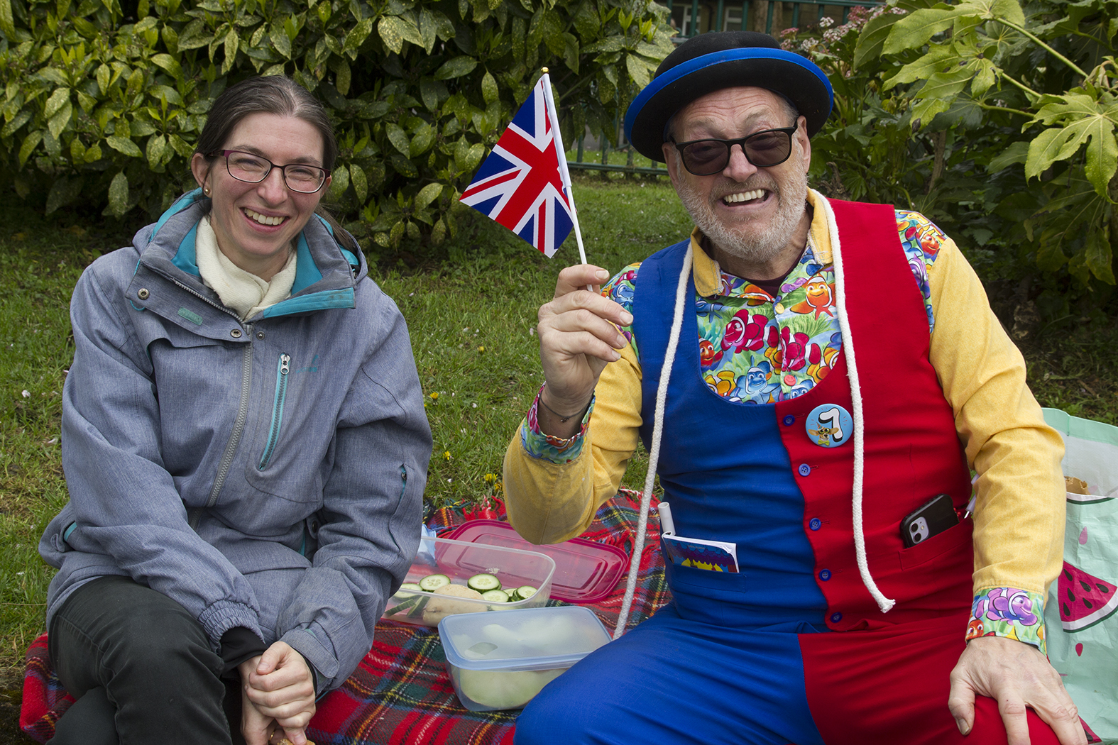 two smiling people one waving a little union jack flag wearing sunglasses and a bowler hat
